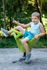 Wall Mural - Little cute boy on green plastic rocking chair in outdoor playground against green summer foliage background