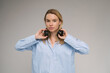 Young Caucasian blonde woman looks at the camera with serious concentration. holding headphones on her neck. Blue casual shirt. Grey background studio shot music audio theme