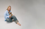 Fototapeta Konie - Smiling blonde woman in a blue shirt sits on the floor, the dog sitting on her laps. Funny cute friends studio portrait. Grey background. Large horizontal size banner copy empty space