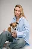 Fototapeta Konie - Smiling blonde woman in a blue shirt sits on the floor, the dog sitting on her laps. Funny cute friends studio portrait. Grey background. Vertical composition