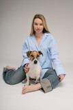 Fototapeta Konie - Woman and dog happy friends sitting on the floor looking at camera and smiling. Professional photo studio. Pretty blonde woman in blue jeans shirt and pet Jack Russell terrier. Vertical composition