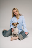 Fototapeta Konie - Woman and dog friends sitting on the floor looking at camera and smiling. Professional photo studio. Pretty blonde woman in blue clothes and pet Jack Russell terrier. Vertical composition