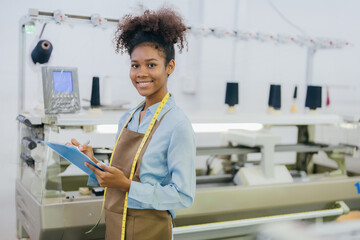 Wall Mural - American female designer has a happy smile holding a list of notes to design clothes, next to which there is a working weaving machine and spools of thread used as materials for production.