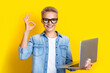 Photo portrait of pretty young girl specs hold netbook show okey symbol wear trendy jeans outfit isolated on yellow color background