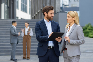 Poster - Two corporate professionals in suits engaged in a discussion with a clipboard outside a modern office building.