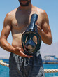 A caucasian man shirtless holds black snorkel mask in his hand against sky, rope railing and blue sea. Snorkeling full face mask diving with a black rim and clear faceplate in a mans hand for swimming