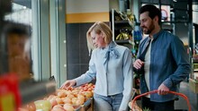 Family Standing Near Display Of Fruits In Supermarket. Attractive Wife Holding Orange While Handsome Husband Carrying Red Basket With Other Products. Concept Of Healthy Eating And Time Together.