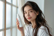 © KP - Radiant Beauty: An attractive Asian woman, illuminated by soft natural light, exudes confidence while holding a cosmetic spray, symbolizing freshness and self-care