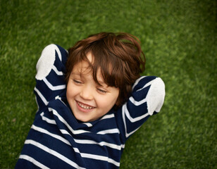 Poster - Child, relax and happy on green grass for holiday, vacation and outdoor wellness in nature. High angle or above of a young kid, boy or student on lawn with excited face for playground, park or spring