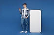 © ViDi Studio - Full body young happy man he wear shirt white t-shirt casual clothes stand near big huge blank screen mobile cell phone with area using smartphone isolated on plain blue background. Lifestyle concept.