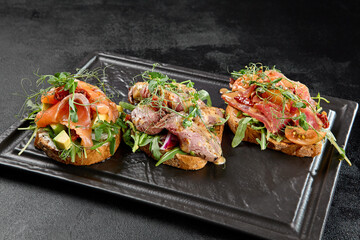 Wall Mural - Assorted bruschetta in black concrete background. Antipasti set: tuna, salmon and roastbeef bruschetta on stone plate. Fish bruschetta and meat bruschetta in minimal style.