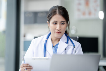 Wall Mural - Portrait of mix race asian middle age female doctor in white lab coat and stethoscope while consult online in laptop. advice on good mental health management and medical treatment costs.