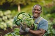 A smiling black working man carrying cabbages in his hands while wearing work gloves in the farm with green big bushes and trees and clear white sky in the background