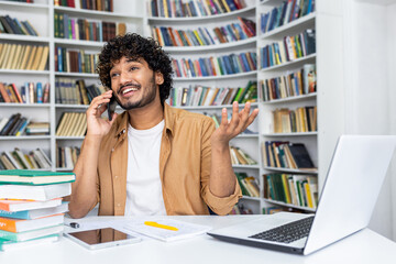 Wall Mural - Happy young man with curly hair makes a phone call while working on his laptop in a home office with bookshelves in the background.