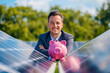 A businessman next to solar panels with a pink piggy bank on them, suggesting savings when using solar panels. Concept of green technology and money savings