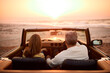 Rear View Of Retired Senior Couple On Vacation In Classic Sports Car At Beach Watching Sunrise
