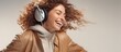 A carefree woman is giggling and smiling, listening to music on headphones while dressed in a stylish tan jacket