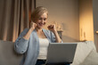 © Grustock - A joyful woman is seated on a sofa with a laptop on her knees, clapping her hands with an expression of elation. The warm, comfortable ambiance of the living room suggests a personal triumph