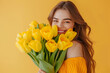 © Baranovsky - Beautiful young woman smiles broadly, her eyes sparkling with joy as she embraces vibrant bouquet of yellow tulips against a sunny backdrop. Mothers day