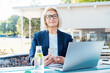 © okrasiuk - Smiling 50's stylish, confident mature business woman, middle aged lady using wireless ear buds, phone and laptop, working remotely in park. Caucasian woman make a video call, study, work online