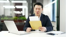 Worried Asian Businessman Reading Letter With Bad News Sitting At Desk At Workplace In Business Office. Disappointed Upset Entrepreneur In Formal Suit Frustrated By Received Unpleasant Notification