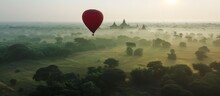 A Red Hot Air Balloon Is Flying Over A Lush Green Field. The Sky Is Hazy And The Sun Is Setting