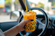 A womans hand holding up a mango boba tea in the car. Orange Bubble tea in the plastic cup with straw.