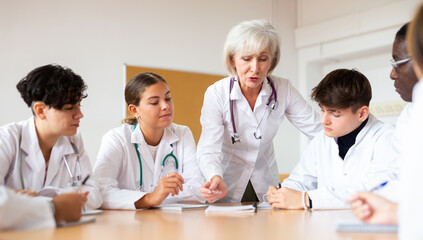 Canvas Print - Group of medical workers in white coats sitting around desk during professional training course, listening to experienced aged female lecturer