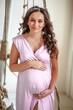 Portrait of a beautiful young pregnant woman in a pink dress. A beautiful pregnant woman in a bright room hugs her stomach with her hands