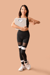 Poster - Beautiful young Asian woman pointing at scales on beige background. Weight loss concept