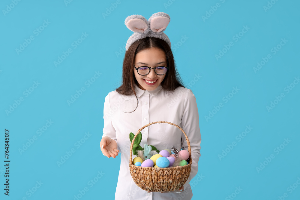 Young Asian businesswoman in bunny ears with Easter eggs on blue background