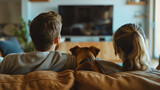 Fototapeta  - Rearview of man and woman, young couple sitting together on sofa with their dog pet and watching TV in living room interior. Family house or home indoors relaxation on couch in an apartment, cuddle