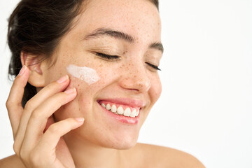 Beauty shot of happy freckled Latin girl model applying face sunscreen or moisturizer cream for perfect hydrated healthy fresh skin care isolated on white background, skincare treatment, close up.