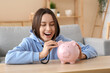 © Pixel-Shot - Cheerful young woman with stethoscope and piggy bank at home. Health insurance concept