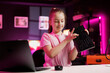 Happy charismatic girl presenting latest gaming computer peripheral tech on her internet channel in neon lit apartment. Trending influencer kid filming review of wireless Bluetooth mechanical keyboard