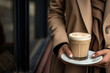Unrecognizable woman in beige coat holding a saucer with a glass of coffee latte outdoors