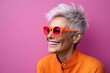 Portrait of a beautiful middle-aged woman with short gray hair wearing orange sunglasses on a pink background
