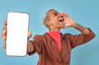 Young inspired African American woman demonstrates phone screen and shouts putting hand to mouth announcing start of sales of new smartphone model stands in turquoise studio. White display, copy space