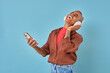 Young cheerful African American woman laughs listening to humorous audio podcast with stand-up stars on headphones and holds phone in hands dressed in casual style stands on turquoise background.