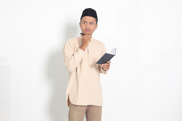 Wall Mural - Portrait of confused Asian muslim man in koko shirt with peci reading a book, thinking about an idea with hand holding pen on chin, looking away. Isolated image on white background