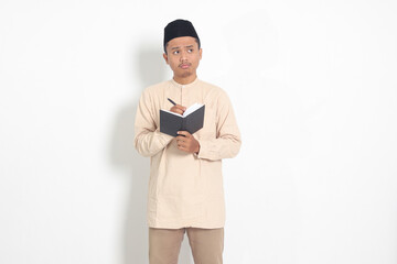 Portrait of confused Asian muslim man in koko shirt with peci reading a book, thinking about an idea with hand holding pen on chin, looking away. Isolated image on white background