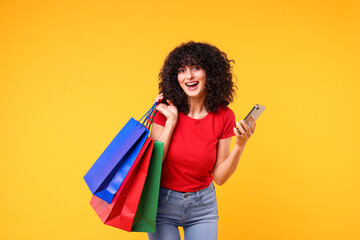 Wall Mural - Happy young woman with shopping bags and modern smartphone on yellow background