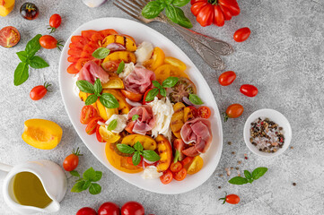 Wall Mural - Caprese salad with mozzarella, tomatoes, prosciutto and grilled peaches on a white plate on a gray concrete background. Fresh summer salad. Top view.