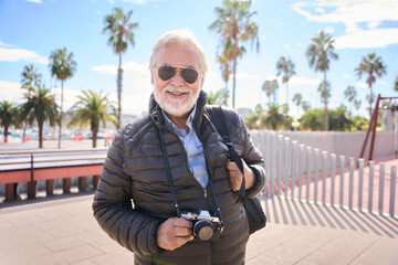 Wall Mural - Portrait of Caucasian mature man tourist with beard and white hair happy and smiling looking at camera enjoying their holidays wearing sunglasses outdoor on a winter sunny day. Copy space