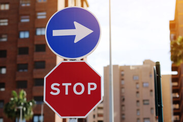 Traffic rules. Red road sign stop and blue and white right directional arrow on residential buildings background. Street sign direction pointing turn right, but need to stop before. Two road signs.