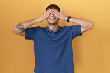 Canvas Print - Young hispanic man standing over yellow background covering eyes with hands smiling cheerful and funny. blind concept.