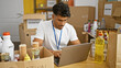 © Krakenimages.com - A focused hispanic man with a beard volunteers at a food donation center, managing inventory on a laptop.