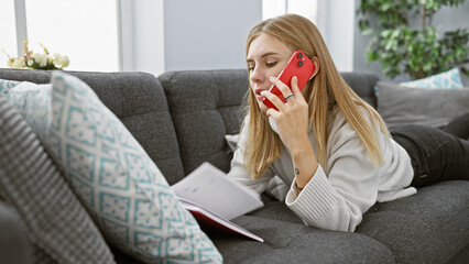 Sticker - Blonde woman conversing on red smartphone lying comfortably on couch in modern living room setting