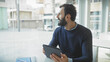 © Krakenimages.com - Bearded man looks pensively out of a window in an office, holding a tablet, with notebook and urban background visible.