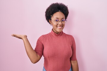 Poster - Beautiful african woman with curly hair standing over pink background smiling cheerful presenting and pointing with palm of hand looking at the camera.